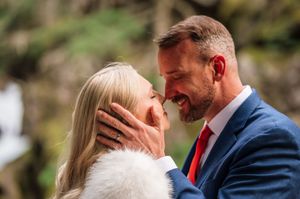 Couple sharing a romantic moment, groom in blue suit and red tie, bride in white fur wrap, outdoors.