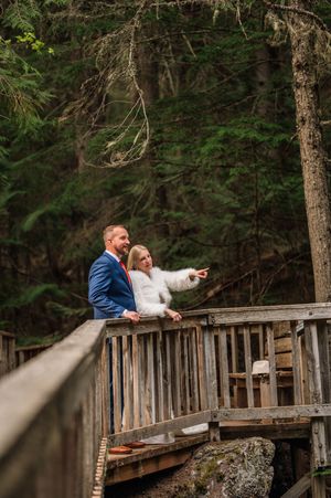 Bride and groom stand on rustic wooden bridge in forest, bride pointing into distance during wedding photos.