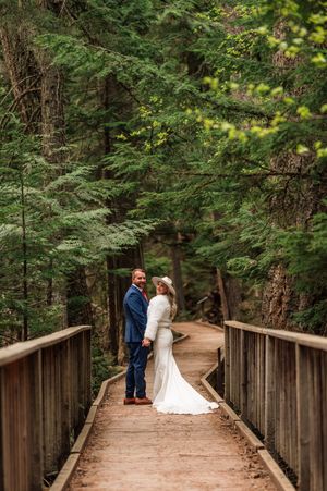Wedding couple holding hands on wooden bridge surrounded by lush green forest trees.