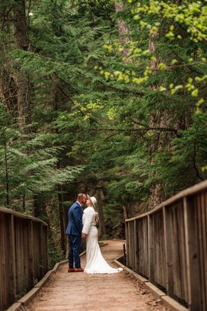 Bride and groom sharing a kiss on a wooden bridge pathway through a dense green forest.