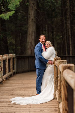 Bride and groom embrace on wooden bridge surrounded by tall evergreen trees in elegant wedding attire.