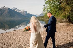 Bride and groom walking along a rocky lakeside shore with snow-capped mountains reflected in calm water.