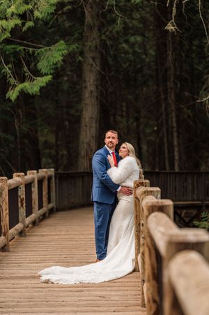 Bride and groom embrace on a wooden bridge surrounded by tall evergreen trees in a romantic winter wedding photo.