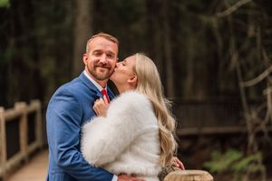 Bride in white fur wrap kisses groom in blue suit on cheek outdoors near wooden bridge in forest.