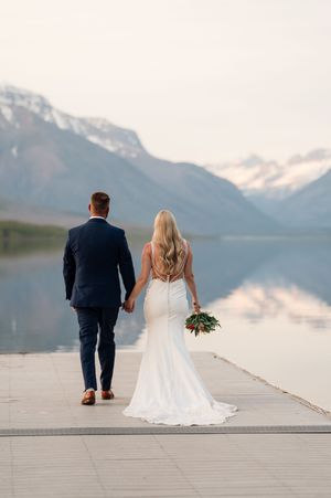 Bride and groom walking hand in hand on dock with mountain lake reflection backdrop.