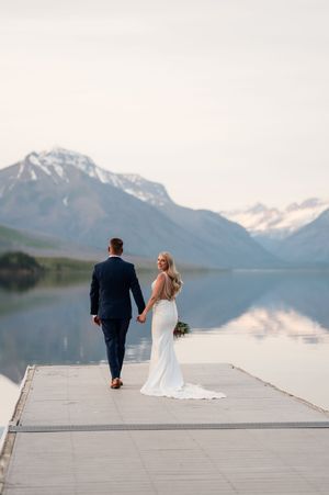 Bride and groom hold hands walking on a dock with stunning mountain lake reflections at sunset.