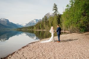 Bride and groom holding hands walking along scenic lake shore with snow-capped mountains at Glacier National Park.