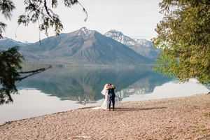 Wedding couple embracing on rocky lakeshore with stunning mountain reflections in calm water.