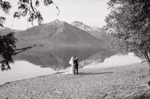Black and white photo of bride and groom kissing on rocky lakeshore with mountain reflections behind them.