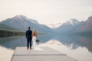 Bride and groom stand on a dock overlooking a serene mountain lake with snow-capped peaks at sunset.