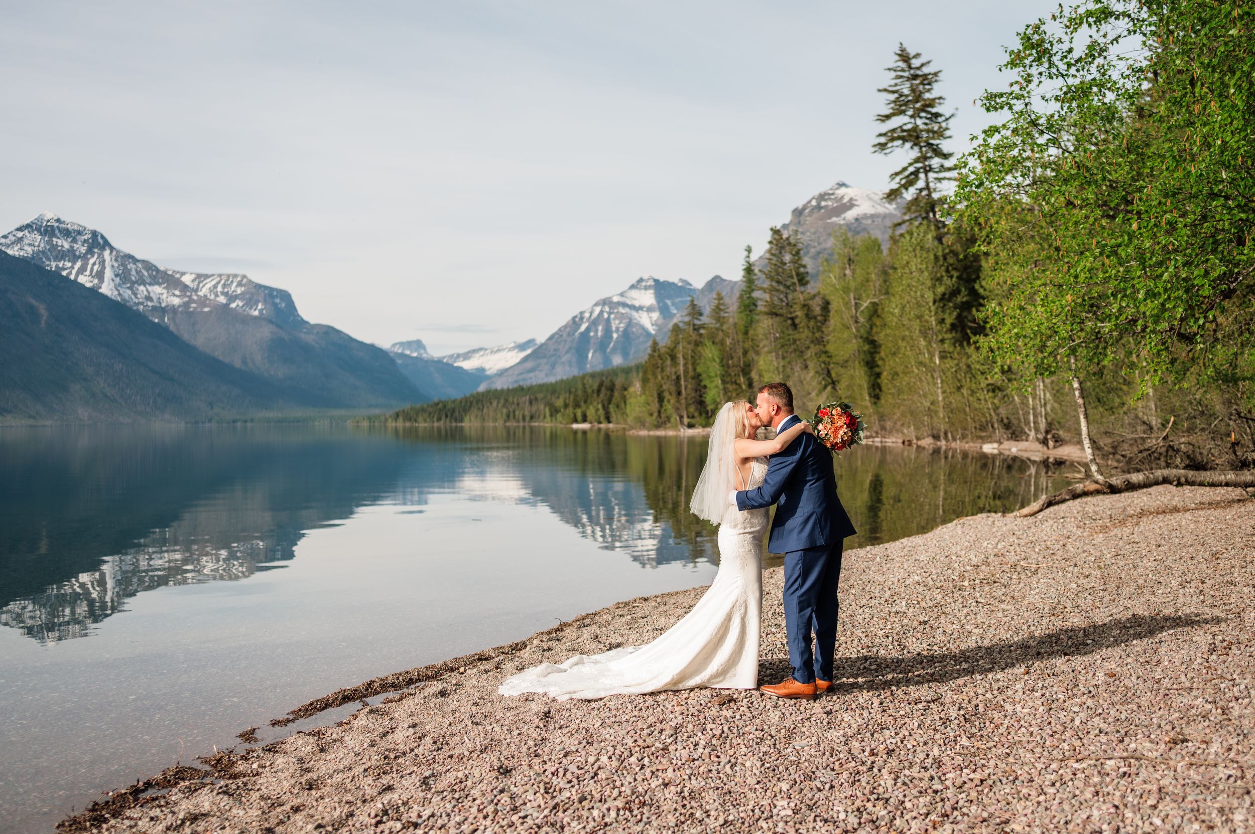 Newlyweds sharing romantic embrace on pebble beach with breathtaking mountain lake reflections at Glacier National Park.