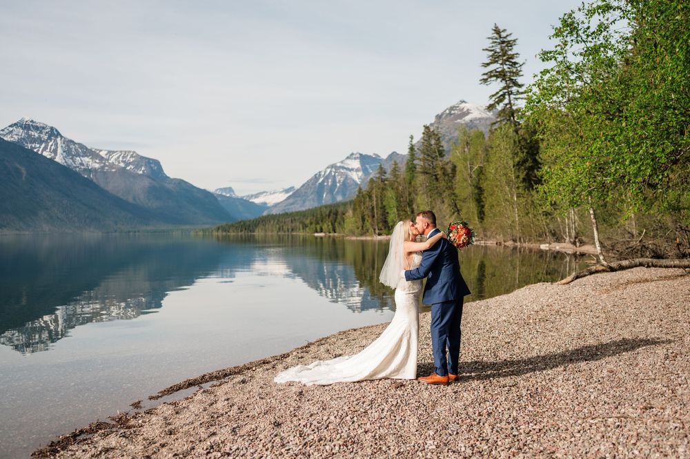 Newlyweds sharing romantic embrace on pebble beach with breathtaking mountain lake reflections at Glacier National Park.