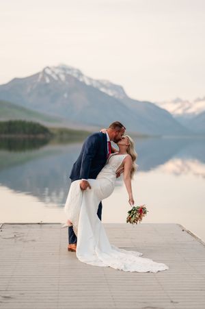 Groom dips bride for a romantic kiss on a dock with snow-capped mountains reflected in calm lake.