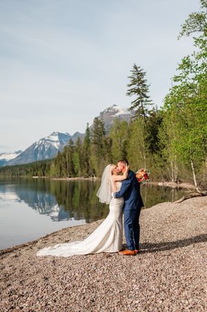 Bride and groom share a kiss on a rocky lakeside with mountain and pine tree reflections in calm water.