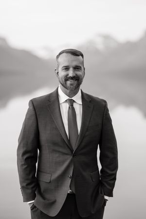 Handsome bearded groom in navy suit smiling confidently with misty mountain lake backdrop, black and white portrait.