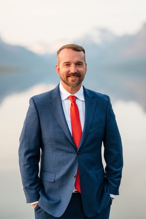 Man in navy suit and red tie smiling outdoors with misty mountain lake backdrop.