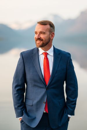 Smiling groom in navy suit and red tie standing before misty mountain lake backdrop.