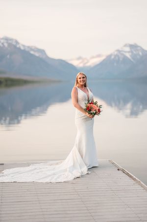 Bride in white gown holding bouquet on dock with mountain lake reflection behind her.