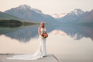 Bride in fitted white gown holding bouquet on dock with snow-capped mountain lake view.