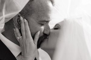 Black and white photo of bride and groom sharing a romantic kiss under a flowing wedding veil.