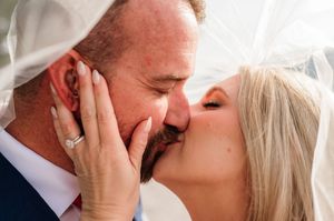 Bride and groom share a romantic kiss under a wedding veil, bride wearing diamond engagement ring.