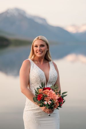 Radiant blonde bride in lace wedding gown holding coral and pink bouquet with mountain lake reflection behind her.