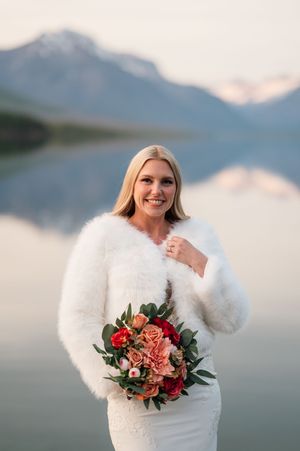 Smiling blonde bride in white fur wrap holds a vibrant red and pink bouquet with mountain lake backdrop at golden hour.