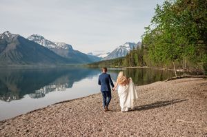 Bride and groom walk hand in hand along rocky lakeshore with stunning snow-capped mountain reflections.