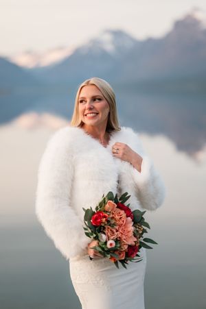Smiling bride in white lace gown and fluffy fur wrap holds colorful bouquet before misty mountain lake.
