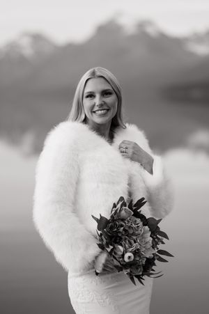 Smiling bride in a white fur jacket holds a bouquet with snowy mountains reflected in a lake behind her.