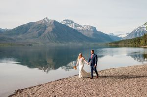 Bride and groom hold hands walking along a rocky lakeshore with snow-capped mountains reflecting in calm water.