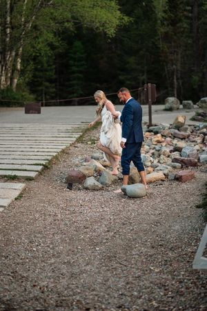 Bride and groom walk barefoot along a rocky shoreline path surrounded by lush forest.