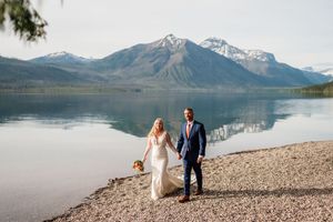 Bride and groom hold hands on a pebble beach with breathtaking snow-capped mountain lake backdrop.