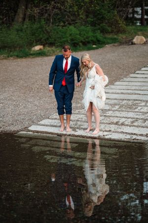 Bride and groom walk barefoot on stone path near water, man in blue suit red tie, woman in white dress and fur wrap.