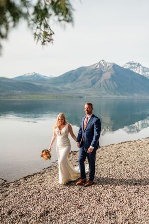 Bride holding colorful bouquet and groom in navy suit pose by a serene mountain lake shore.