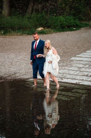 Bride and groom wade through shallow water in wedding attire with forest reflections below.
