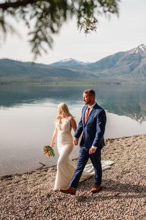 Bride and groom walking hand in hand along rocky lakeside with snow-capped mountains.