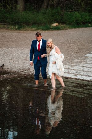 Bride and groom wading barefoot in shallow water on rocky beach, laughing together.
