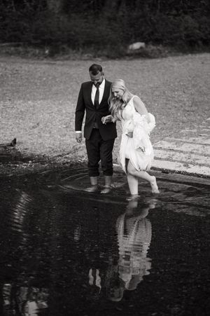 Bride and groom wade through shallow water in black and white wedding photo with reflections.