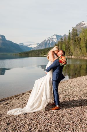 Bride and groom embrace on rocky lakeshore with snow-capped mountains reflected in calm water during wedding photos.