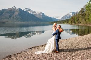 Groom lifts bride on rocky lakeshore with breathtaking snow-capped mountains and calm lake reflection.