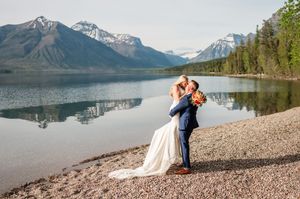 Groom lifts bride in a romantic embrace on a rocky lakeshore with stunning mountain scenery.