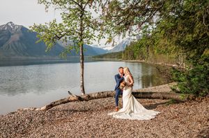 Bride and groom share a tender moment under a lakeside tree with majestic mountain views.
