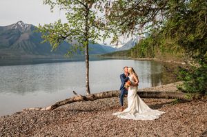 Bride and groom embrace near fallen log by mountain lake with scenic peaks and trees backdrop.