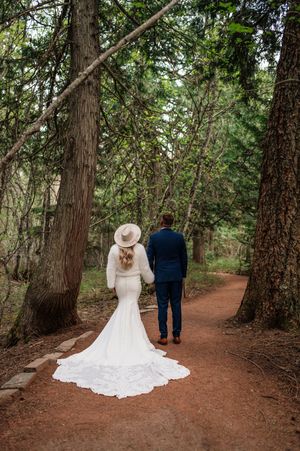 Bride and groom walking away on forest path, her white gown trailing behind them.