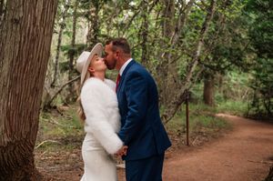 Bride and groom kiss on a forest path, bride in white fur wrap and hat, groom in navy suit.