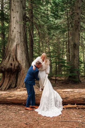Groom in navy suit lifts bride's lace train as she laughs among towering evergreen trees in forest.