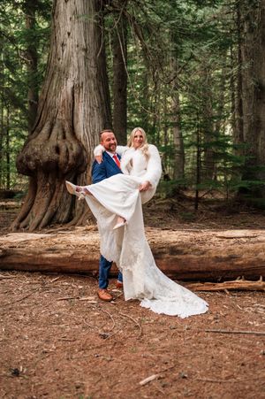 Groom lifting bride in white gown and fur wrap beside large tree in forest.