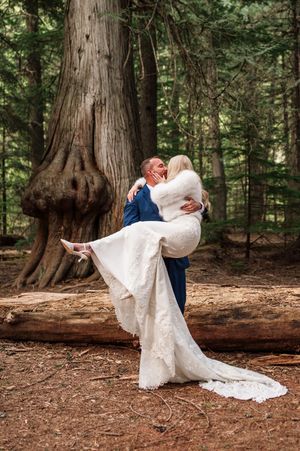 Groom in navy suit lifts bride in lace gown for a kiss beside a massive tree in pine forest.