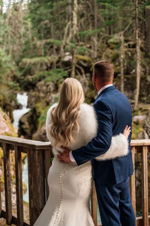 Bride and groom embrace on a wooden overlook viewing a forest waterfall, bride wearing a fur wrap.